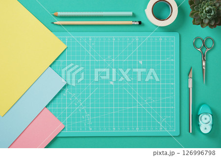 Top view of a desk for paper crafts. Crafting tools. Turquoise cutting mat, scissors, craft knife, pencil, glue, tape, colored paper. Space for your text. 126996798