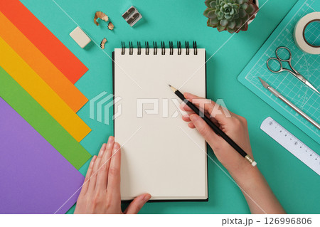 A person is preparing to draw on a spiral-bound notebook with a black pencil, surrounded by colorful sheets of paper and craft tools. Space for your text or drawing A person is preparing to draw on a spiral-bound notebook with a black pencil, surrounded by colorful sheets of paper and craft tools. Space for your text or drawing 126996806