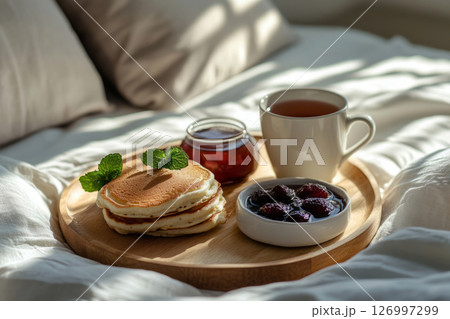 A well-prepared breakfast board featuring fluffy pancakes topped with cream and mint, alongside a cup of warm tea, enjoyed in a serene bedroom adorned with natural light 126997299