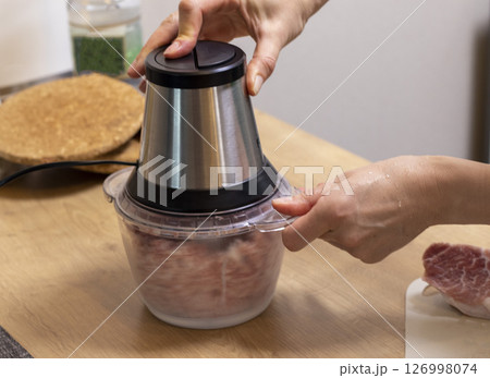 Woman mince the meat in the electrical chopper on the kitchen counter top 126998074