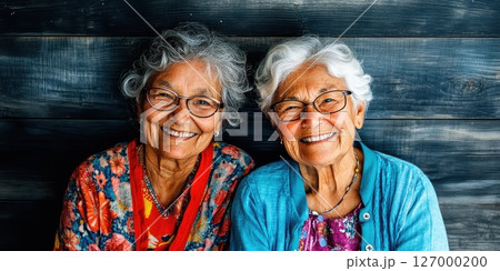 Smiling elderly women share a joyful moment together, sitting against a simple wooden backdrop in the afternoon sunlight, showcasing the beauty of friendship and laughter 127000200
