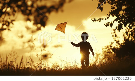 Child flying a kite at sunset in a grassy field with golden hues illuminating the sky and creating a serene atmosphere perfect for outdoor activities 127001229