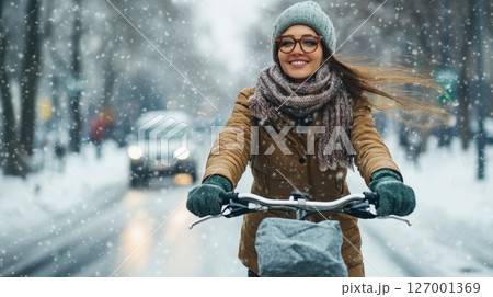 Joyful woman cycling in snow-covered city street during winter season while wearing warm clothing and enjoying the snowfall 127001369