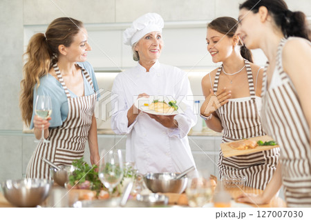 Senior woman host of cooking courses shows visitors of workshop cooked chicken under creamy sauce Senior woman host of cooking courses shows visitors of workshop cooked chicken under creamy sauce 127007030