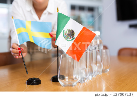 A woman in a white blouse with friendly smile arrangs flags of Mexico and Sweden for meeting. A woman in a white blouse with friendly smile arrangs flags of Mexico and Sweden for meeting. 127007137