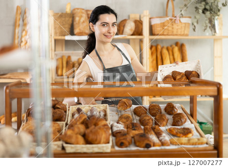 Young woman employee puts croissants in window, arranges display of goods at bakery. 127007277