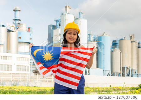 Positive girl in overalls with Malaysia flag in her hands against the background of chimneys of modern factory 127007306