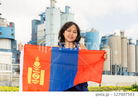 Portrait of positive girl in construction overalls with the Mongolia flag on the background of modern factory Portrait of positive girl in construction overalls with the Mongolia flag on the background of modern factory 127007503