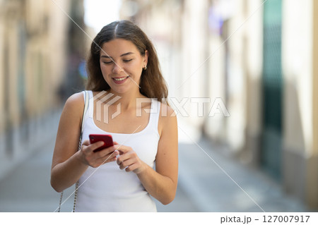 Happy woman looking to phone in hands and chatting in city Happy woman looking to phone in hands and chatting in city 127007917