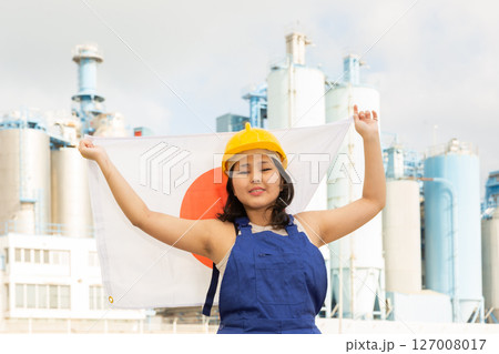 Happy young woman in helmet holding flag of Japan against background of factory 127008017