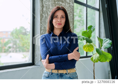 Portrait of beautiful confident serious woman looking at camera with crossed arms, near window Portrait of beautiful confident serious woman looking at camera with crossed arms, near window 127008116