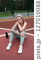 The concept of energy, vitality, well-being. A toned, beautiful blonde in sports clothes is relaxing after a workout outdoors. She's sitting on a sports basketball court 127010088