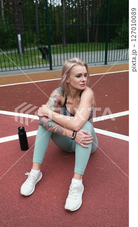 The concept of energy, vitality, well-being. A toned, beautiful blonde in sports clothes is relaxing after a workout outdoors. She's sitting on a sports basketball court The concept of energy, vitality, well-being. A toned, beautiful blonde in sports clothes is relaxing after a workout outdoors. She's sitting on a sports basketball court 127010089