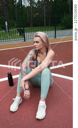 The concept of energy, vitality, well-being. A toned, beautiful blonde in sports clothes is relaxing after a workout outdoors. She's sitting on a sports basketball court The concept of energy, vitality, well-being. A toned, beautiful blonde in sports clothes is relaxing after a workout outdoors. She's sitting on a sports basketball court 127010090