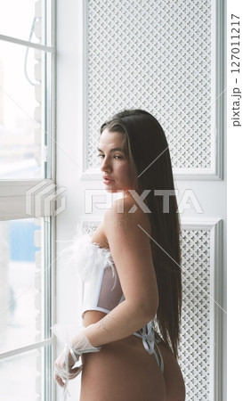 Portrait of young beautiful model posing in white lace lingerie, corset and stockings near a large window. Calm cute brunette with long hair in fashionable lingerie. 127011217
