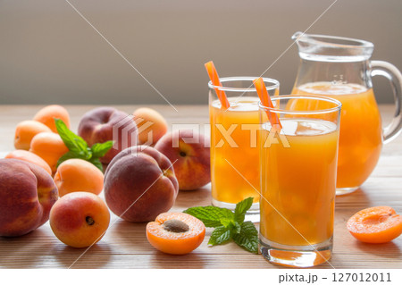 Apricot and peach juice in glasses with ice near the jar of juice and scattered apricots and peaches, mint on a light wooden background. Apricot and peach juice with ice. Horizontal. Daylight. Close. Apricot and peach juice in glasses with ice near the jar of juice and scattered apricots and peaches, mint on a light wooden background. Apricot and peach juice with ice. Horizontal. Daylight. Close. 127012011