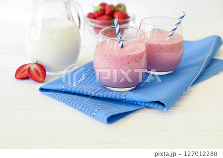 Two colorful strawberry smoothies in clear glasses are placed next to a pitcher of milk and a bowl of fresh strawberries on a blue napkin. Two colorful strawberry smoothies in clear glasses are placed next to a pitcher of milk and a bowl of fresh strawberries on a blue napkin. 127012280