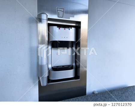 Close up of stainless steel water dispenser with integrated paper cup holder in a public transport cabin. Practical hydration solution for modern urban infrastructure Close up of stainless steel water dispenser with integrated paper cup holder in a public transport cabin. Practical hydration solution for modern urban infrastructure 127012370