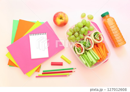 School supplies and lunch box with tasty rolls, cucumbers, carrots, grapes, apple, bottle of juice on light pink background, back to school concept. Horizontal. Top view. 127012608
