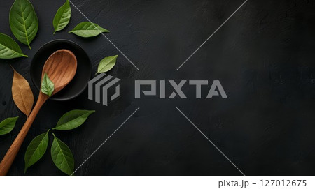 Wooden spoon and green leaves on black background for culinary or nature themes. Minimalist setup with fresh foliage for kitchen or wellness designs. Perfect for organic or rustic concepts. 127012675