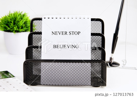 NEVER STOP BELIEVING motivational phrase on a white sheet of paper in a black stand on a white background 127013763