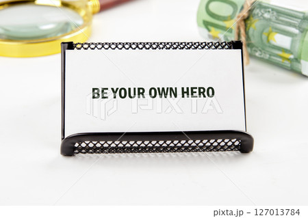 Motivational words BE YOUR OWN HERO on a white business card in a composition with a magnifying glass and banknotes in the background 127013784