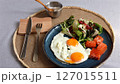 Bright sunny eggs with orange yolks beside green salad, pomegranate seeds, and salmon slices on blue plate, served with silver bowl of salt, emphasizing eco-conscious healthy lifestyle. 127015511