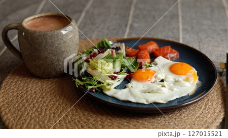Two sunny eggs with orange yolks rest next to salmon slices, fresh salad with pomegranate seeds, and creamy smoothie in gray mug, highlighting eco-conscious healthy lifestyle meal. Two sunny eggs with orange yolks rest next to salmon slices, fresh salad with pomegranate seeds, and creamy smoothie in gray mug, highlighting eco-conscious healthy lifestyle meal. 127015521
