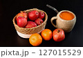 Ceramic mug filled with pumpkin beverage near basket of red apples, oranges, pomegranate on dark surface, vibrant autumn setup focusing on fresh seasonal produce, rustic charm, organic tones. 127015529