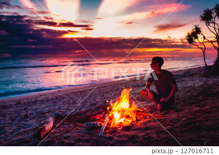 Alone man sitting by a beach bonfire at sunset, enjoying peaceful solitude and stunning tropical ocean view. 127016711