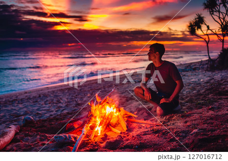Man sit alone by a beach bonfire at sunset, enjoying peaceful solitude and stunning ocean view. Man sit alone by a beach bonfire at sunset, enjoying peaceful solitude and stunning ocean view. 127016712