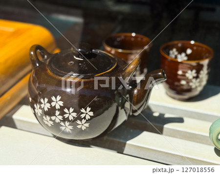 Dark brown teapot with floral pattern. Ceramic teaware set decorated with white cherry blossoms in window display. 127018556