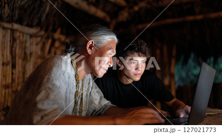 Elder and young programmer work together to create a digital archive of their native language in a shaded hut 127021773