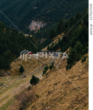Rugged highland path with red car parked at a curve among pine-covered mountain slopes 127022509