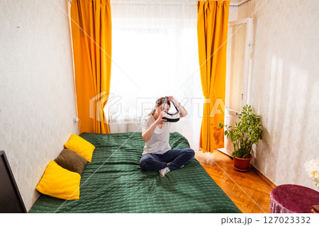 Woman lost in a virtual world while sitting on bed using a VR headset, connecting with immersive AI-driven experiences. Wide shot, top view Woman lost in a virtual world while sitting on bed using a VR headset, connecting with immersive AI-driven experiences. Wide shot, top view 127023332