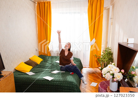 A happy woman scatters bills and payment receipts while sitting at home on her bed. Concept of economy and investments. Wide shot 127023350