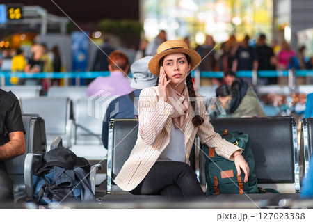 Wide shot of young Caucasian woman in a hat and jacket is sitting in an armchair in the waiting room and talking on a smartphone. Waiting to board the plane Wide shot of young Caucasian woman in a hat and jacket is sitting in an armchair in the waiting room and talking on a smartphone. Waiting to board the plane 127023388