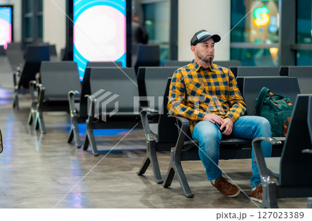 Wide shot of adult Caucasian man in cap and jacket using smartphone in airport waiting area sitting in chair. Concept of travel app Wide shot of adult Caucasian man in cap and jacket using smartphone in airport waiting area sitting in chair. Concept of travel app 127023389