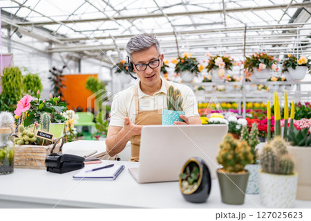 A Caucasian florist assisting a customer online in a greenhouse setting, showcasing a cactus on a video call. 127025623
