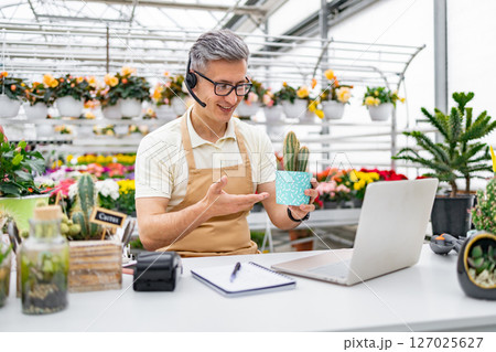 A florist on a video call, showing a cactus to a customer. The shop has a large selection of flowers. A florist on a video call, showing a cactus to a customer. The shop has a large selection of flowers. 127025627