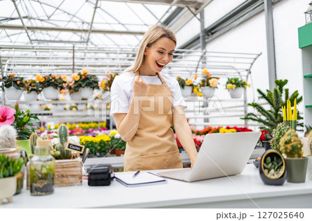 A cheerful Caucasian woman celebrates a successful sale in her flower shop, working at the table, in front of flowers and plants. 127025640