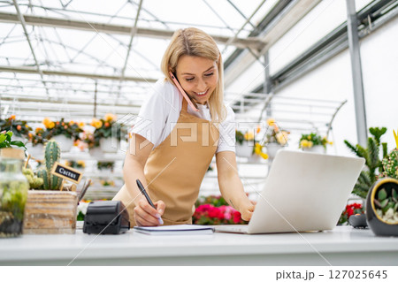 A Caucasian woman working in a florist shop, taking a call and using a laptop while writing. A Caucasian woman working in a florist shop, taking a call and using a laptop while writing. 127025645
