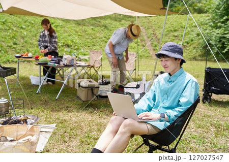 キャンプ中にパソコン作業する男性と食事準備をする仲間たち 127027547