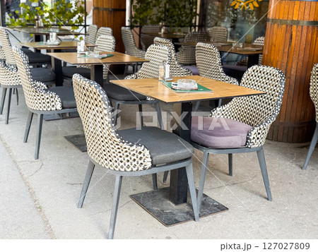 Outdoor restaurant seating with woven chairs and wooden tables. Arranged dining setup with ashtrays and placemats on concrete pavement. Outdoor restaurant seating with woven chairs and wooden tables. Arranged dining setup with ashtrays and placemats on concrete pavement. 127027809