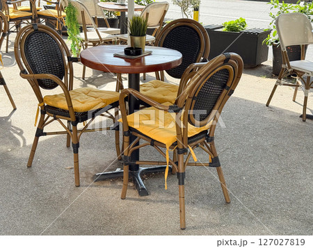 Outdoor cafe seating with yellow cushions. Round wooden table with rattan chairs and potted plant centerpiece. 127027819