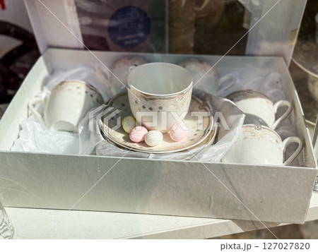 Porcelain tea set with floral border in gift box. Elegant cups and saucers arranged with pastel sweets on satin fabric 127027820