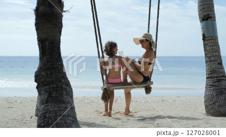 Mom and daughter swing and laugh on sunny tropical beach as girl drinks shake by the sea Mom and daughter swing and laugh on sunny tropical beach as girl drinks shake by the sea 127028001
