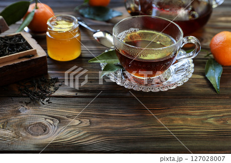 Glass cup holds steaming tea accompanied by small jar of honey and fresh oranges. Dried tea leaves spread on wooden surface create a warm atmosphere perfect for afternoon relaxation. Glass cup holds steaming tea accompanied by small jar of honey and fresh oranges. Dried tea leaves spread on wooden surface create a warm atmosphere perfect for afternoon relaxation. 127028007