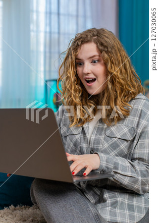 Woman sitting on sofa with laptop raising eyebrows surprised by unexpected good news or discovery 127030065