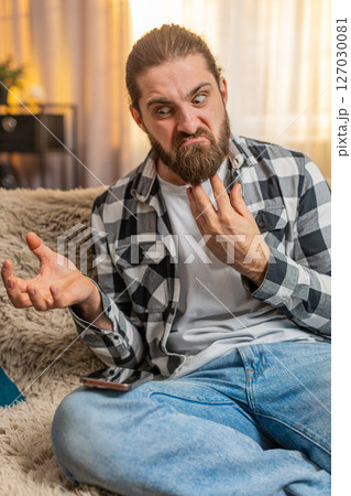 Young man anxious at home, urgent phone call on knee, frustration, loud speaker, intense quarrel Young man anxious at home, urgent phone call on knee, frustration, loud speaker, intense quarrel 127030081